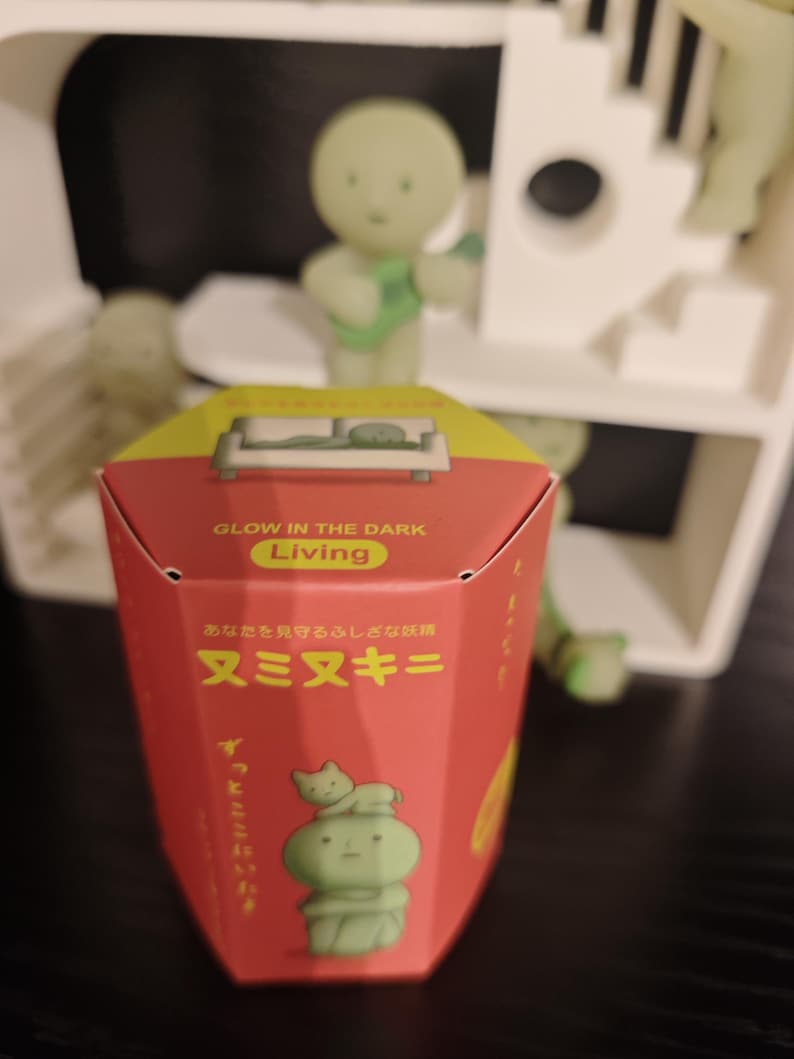 May include: A red box with the text "GLOW IN THE DARK Living" and Japanese characters. The box features a green cartoon figure with a cat on its head. A green figure playing a guitar is in the background.