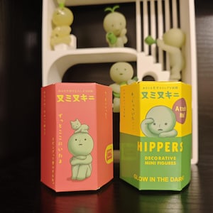 May include: Two decorative mini figure boxes, one red and one yellow-green, with Japanese text and illustrations. The red box features a figure with a cat, while the yellow-green box says "HIPPERS" and "GLOW IN THE DARK". Several small, glowing green figures are displayed on a white shelf.