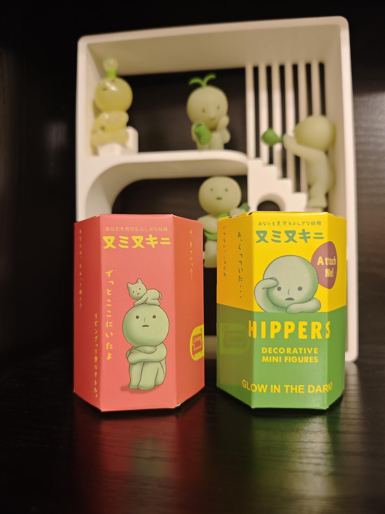May include: Two decorative mini figure boxes, one red and one yellow-green, with Japanese text and illustrations. The red box features a figure with a cat, while the yellow-green box says "HIPPERS" and "GLOW IN THE DARK". Several small, glowing green figures are displayed on a white shelf.