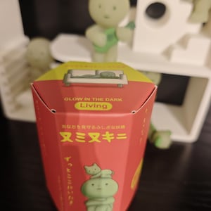 May include: A red box with yellow accents and Japanese text, featuring a glow-in-the-dark theme. The box has an illustration of a green figure with a cat. The text "GLOW IN THE DARK Living" is visible. There are also green figurines in the background.