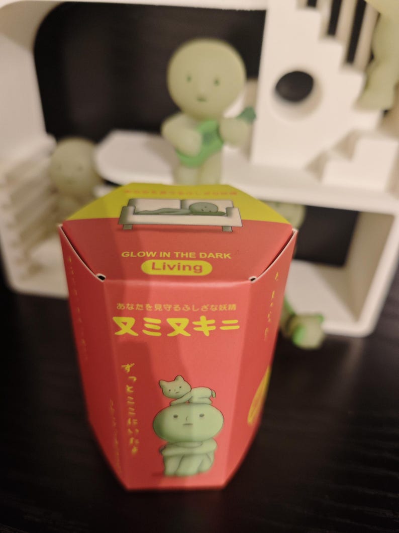 May include: A red box with yellow accents and Japanese text, featuring a glow-in-the-dark theme. The box has an illustration of a green figure with a cat. The text "GLOW IN THE DARK Living" is visible. There are also green figurines in the background.