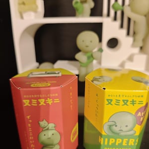 May include: Two hexagonal boxes for decorative mini figures, one red and one yellow, with Japanese text and illustrations of green figures. The boxes are labelled "Glow in the Dark Living" and "Hippers! Decorative Mini Figures". Several glowing green figures are in the background.