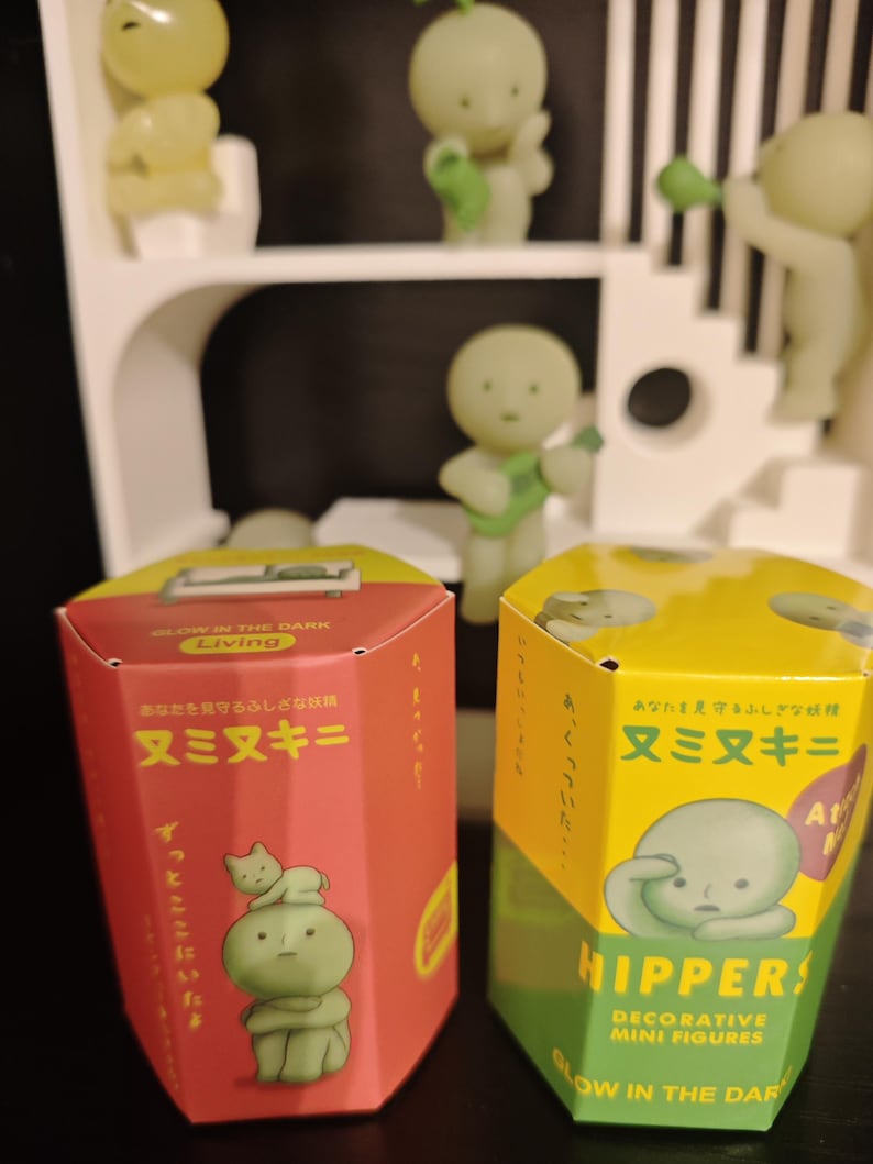 May include: Two hexagonal boxes for decorative mini figures, one red and one yellow, with Japanese text and illustrations of green figures. The boxes are labelled "Glow in the Dark Living" and "Hippers! Decorative Mini Figures". Several glowing green figures are in the background.