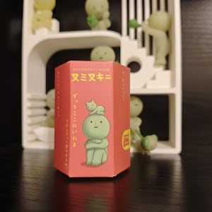 May include: A red hexagonal box with Japanese text and an illustration of a green figure with a cat on its head. The box is in front of a white shelf with several small, green, glowing figures.