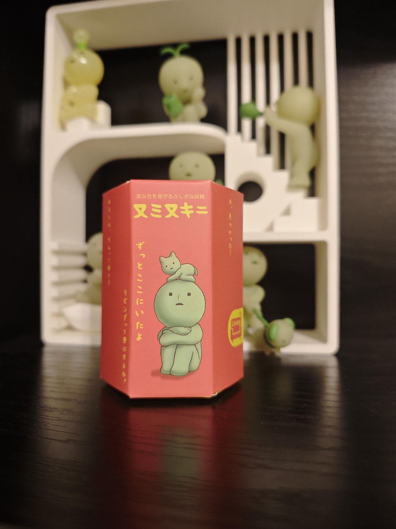 May include: A red hexagonal box with Japanese text and an illustration of a green figure with a cat on its head. The box is in front of a white shelf with several small, green, glowing figures.
