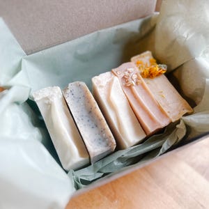 Könnte beinhalten: Eine Geschenkbox mit vier handgemachten Seifen. Die Seifen sind in verschiedenen Farben erhältlich, darunter Weiß, Grau und Rosa, und sind mit Samen und getrockneten Blumen verziert. Die Box ist mit grünem Seidenpapier ausgekleidet.