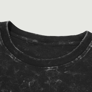 May include: Close-up of a black t-shirt neckline. The shirt has a distressed, faded appearance with a crew neck collar. The fabric has a mottled texture, with lighter areas contrasting against the dark base color.