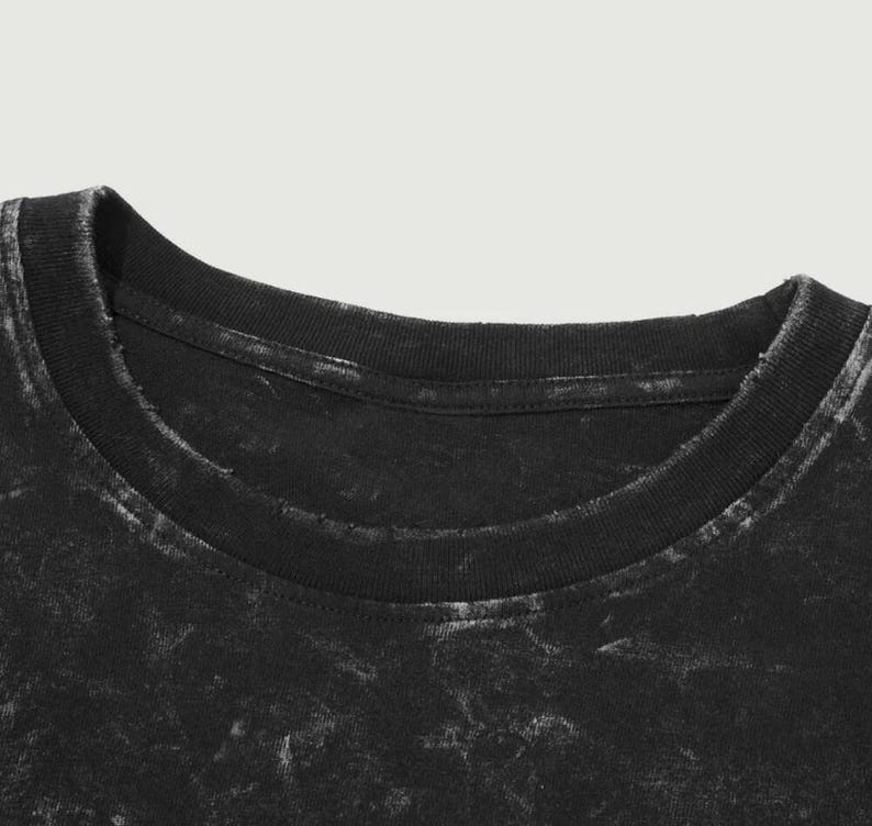 May include: Close-up of a black t-shirt neckline. The shirt has a distressed, faded appearance with a crew neck collar. The fabric has a mottled texture, with lighter areas contrasting against the dark base color.