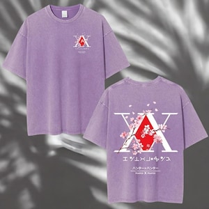 May include: A lilac-colored t-shirt featuring a graphic design on the front and back. The front design includes a red and white emblem with Japanese text. The back design features a large white "X" with a red diamond and cherry blossom accents.