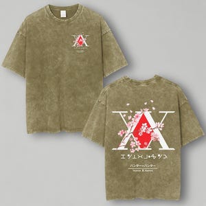May include: Olive green t-shirt featuring a graphic design on the front and back. The front design includes a red and white emblem with Japanese text. The back design features a large white "X" with a red diamond, cherry blossoms, and Japanese text. The text "Hunter x Hunter" is also visible.