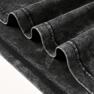 May include: Close-up of a dark gray, acid-washed fabric with visible texture and folds. The fabric has a distressed appearance, with seams and stitching details. The material appears to be a soft, possibly cotton, textile.