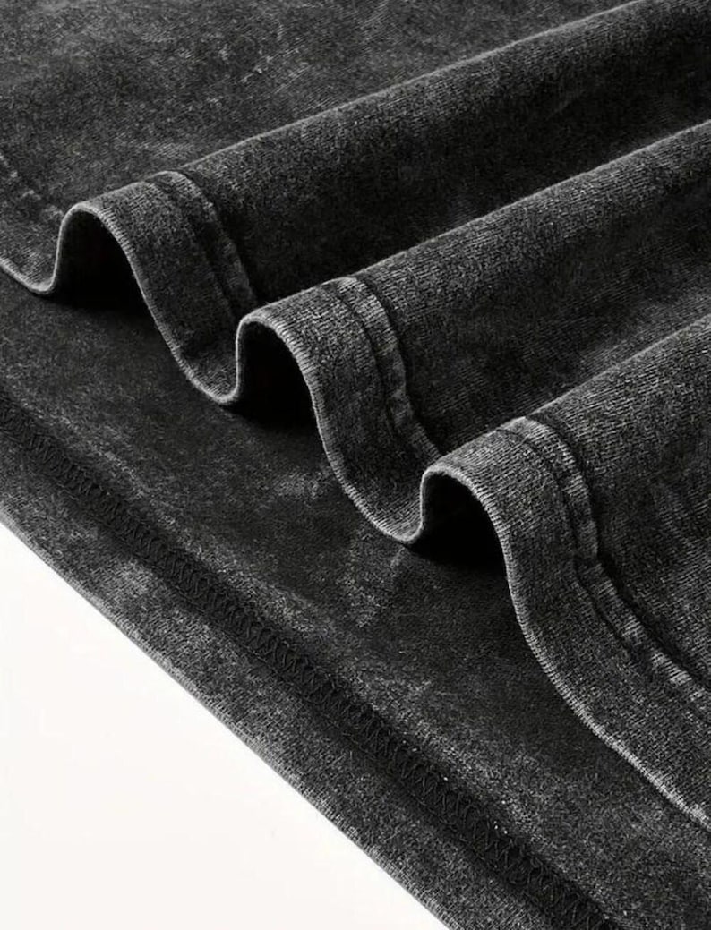 May include: Close-up of a dark gray, acid-washed fabric with visible texture and folds. The fabric has a distressed appearance, with seams and stitching details. The material appears to be a soft, possibly cotton, textile.