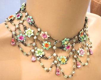 May include: A multi-strand necklace with colourful floral accents. The necklace has a bronze-tone metal base with layers of small, round beads and dangling teardrop-shaped pink crystals. The flowers are in shades of pink, orange, and green.