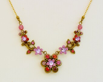Könnte beinhalten: Blumenhalskette mit messingfarbenem Metallrahmen. Die Halskette hat rosa, rote und violette Kristalle in einem Blumen- und Rankenmuster. Die Kette ist goldfarben, und die Halskette ist vor einem neutralen Hintergrund platziert.
