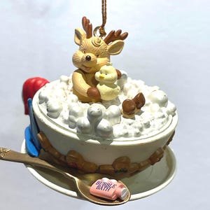 May include: A whimsical Christmas ornament featuring a reindeer and a duck in a bubble bath. The ornament has a gold-colored chain for hanging. A small pink bottle of "Bubble Bath" is on a spoon.