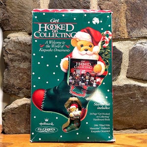 May include: A green and red Hallmark Keepsake Ornament starter set box. The box features a teddy bear wearing a Santa hat and holding a book. The text on the box reads "Get Hooked on Collecting" and "A Welcome to the World of Keepsake Ornaments."