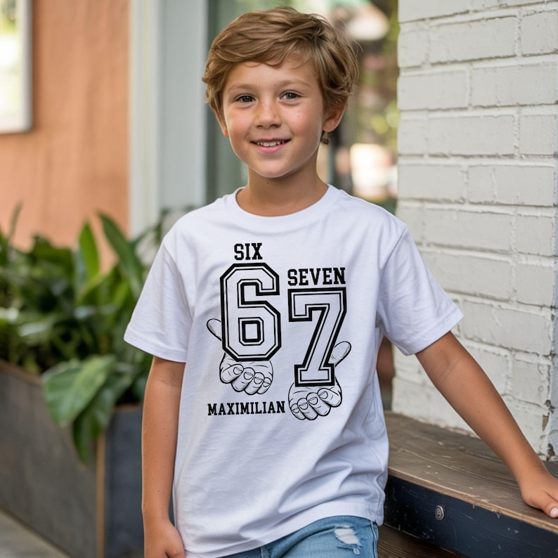 May include: A white short-sleeved t-shirt featuring a black graphic print. The design includes the words SIX SEVEN, the large numbers 67, and the name MAXIMILIAN, accompanied by illustrations of hands making gestures beneath the numbers.