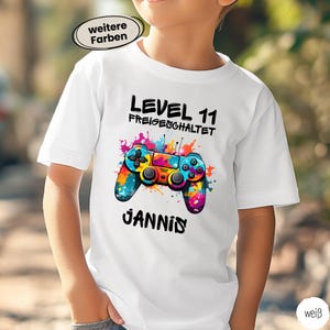 Könnte beinhalten: Weißes T-Shirt mit einem bunten Videospiel-Controller-Motiv. Das Shirt zeigt den Text "LEVEL 11 FREIGESCHALTET" und den Namen "JANNIS" in Schwarz. Das Controller-Design enthält blaue, gelbe und rosa Akzente.