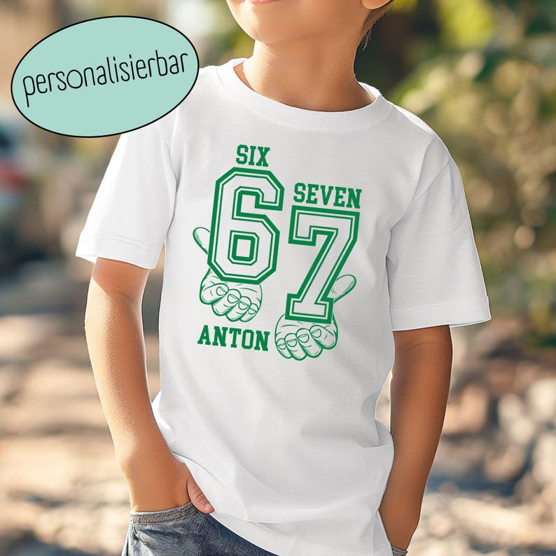 May include: A white short-sleeved t-shirt featuring a green graphic print. The design includes the large numbers 6 and 7, the words SIX, SEVEN, and ANTON, and illustrations of hands forming the numbers with fingers. The text personalisierbar is visible.