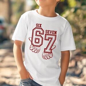 May include: A white short-sleeved t-shirt featuring a graphic print in dark red. The design displays the text SIX SEVEN above the large numbers 67, with stylized illustrations of hands positioned beneath each number to frame the graphic.