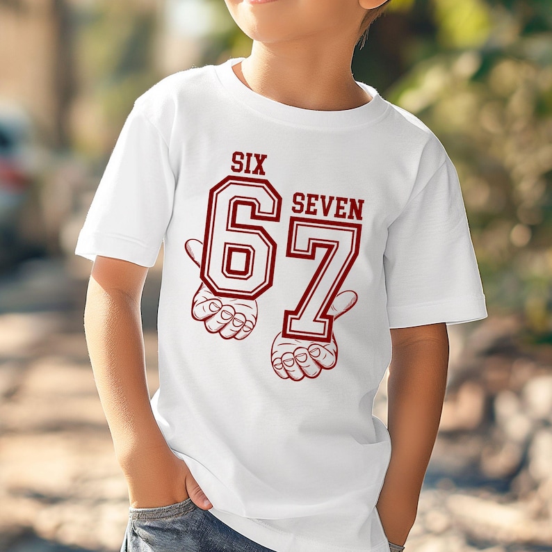 May include: A white short-sleeved t-shirt featuring a graphic print in dark red. The design displays the text SIX SEVEN above the large numbers 67, with stylized illustrations of hands positioned beneath each number to frame the graphic.