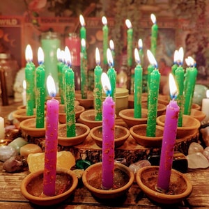 May include: A collection of lit candles in various shades of green and purple, arranged in small, round, terracotta-colored bowls. The candles are on a wooden surface, with a blurred background of foliage and other objects.