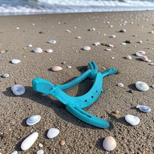 May include: A turquoise horseshoe lies on a sandy beach scattered with seashells. The horseshoe is made of metal and has a slightly weathered appearance. The ocean waves are visible in the background under a clear blue sky.