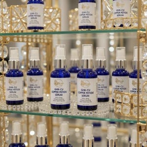 Puede incluir: M&uacute;ltiples frascos de vidrio azul cobalto con dispensadores de bomba blancos y etiquetas que dicen GHK-CU COPPER PETIDE SERUM. Los frascos est&aacute;n dispuestos en estantes de vidrio dentro de un expositor de metal dorado con cristales.