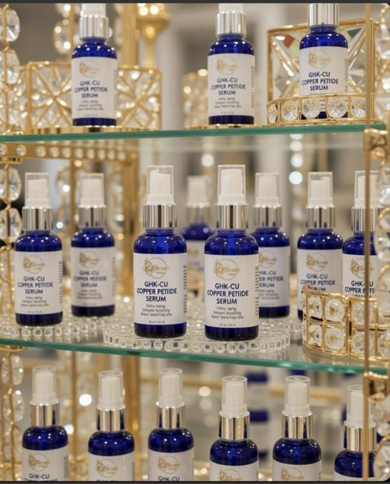 Puede incluir: M&uacute;ltiples frascos de vidrio azul cobalto con dispensadores de bomba blancos y etiquetas que dicen GHK-CU COPPER PETIDE SERUM. Los frascos est&aacute;n dispuestos en estantes de vidrio dentro de un expositor de metal dorado con cristales.