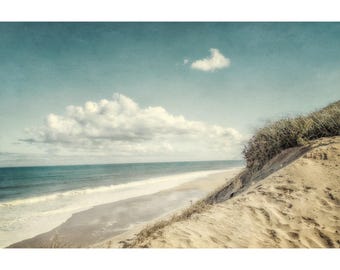 May include: A serene beach scene with a light blue sky and fluffy white clouds. The ocean waves gently roll onto the sandy shore. A sand dune with dry grass sits on the right side of the image, creating a peaceful coastal landscape.
