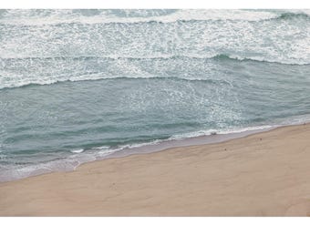May include: An aerial view of a sandy beach with foamy waves crashing on the shore. The water is a light blue color and the sand is a light brown color.