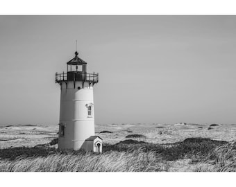 May include: Black and white photograph of a lighthouse on a sandy dune. The lighthouse is white with a black top and is surrounded by tall grass and sand. The sky is clear and cloudless.