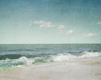 May include: A vintage-style photograph of a sandy beach with turquoise water and a blue sky with white clouds.