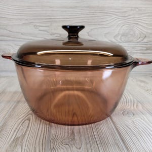 May include: A vintage brown glass casserole dish with a matching lid. The dish has two handles and a dark brown knob on the lid. The glass has a transparent quality, allowing visibility of the contents. A classic kitchenware item.