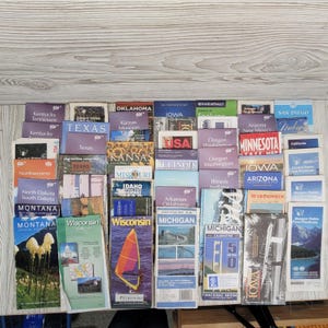May include: A collection of vintage state highway maps, including Texas, Montana, and Wisconsin. The maps are in various sizes and colors, with state names prominently displayed. The maps are arranged on a light-colored wooden surface.