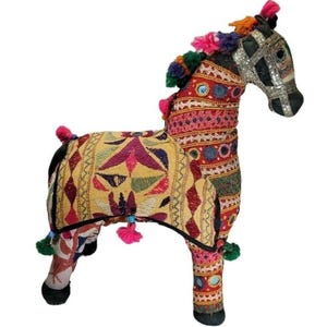 May include: A decorative stuffed horse figurine with colorful embroidered fabric. The horse features a yellow saddle blanket with geometric patterns, a red and pink body, and a black head with silver accents. It is adorned with tassels and colorful details.