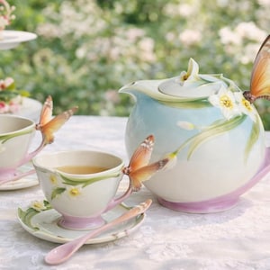 May include: A porcelain tea set with a teapot and two teacups, each adorned with butterfly handles and floral accents. The teapot is light blue with a pink base and handle. The teacups have matching saucers and pink spoons.
