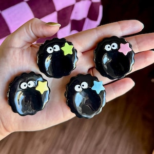 May include: Five black, round, glossy pins with white eyes and colorful star accents. The stars are in shades of green, pink, and blue. The pins are held in a hand against a blurred background.
