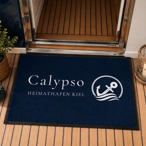 Könnte beinhalten: Eine rechteckige, marineblaue Fußmatte mit dem weißen Schriftzug Calypso und HEIMATHAFEN KIEL sowie einer weißen Grafik eines Ankers, der von einem Seil und stilisierten Wellen umgeben ist, auf einem Holzboden.