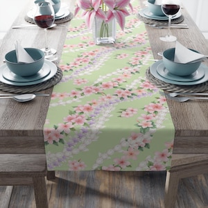 May include: A pale green table runner featuring a floral design of pink, white, and lilac flowers. The runner is laid on a wooden table, set with plates, bowls, and glasses for a meal.