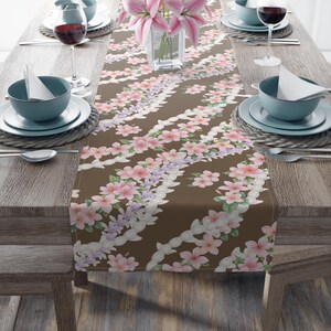 May include: A brown floral table runner with pink and purple flowers and white detailing. The table is set with blue bowls, white plates, wine glasses, and cutlery. A vase of pink lilies sits in the centre of the table.