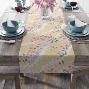 May include: A floral table runner with a pale yellow backdrop. The runner displays a repeating pattern of pink, purple, and white flowers. The table is set with blue bowls, white plates, and wine glasses.