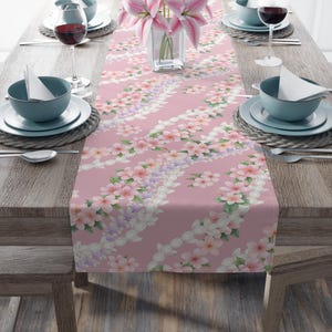 May include: A pink floral table runner featuring a repeating pattern of white, pink, and purple flowers. The runner is placed on a wooden table with place settings, including plates, bowls, and wine glasses. A vase of pink lilies is in the centre.