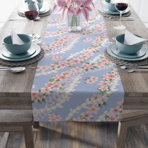 May include: A pale blue table runner adorned with a floral design. The runner showcases rows of pink, white, and lilac flowers. The table is set with blue bowls, plates, and wine glasses. A vase of pink lilies is in the centre.