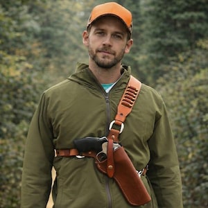 Könnte beinhalten: Ein braunes Leder-Schulterholster mit einer schwarzen Handfeuerwaffe und einem Gürtel mit Patronen. Das Holster wird über einer grünen Jacke und einer braunen und orangefarbenen Kappe getragen.