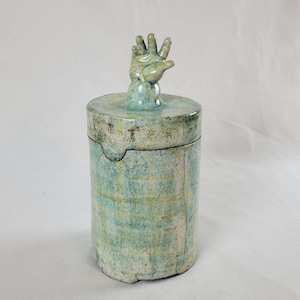 May include: A cylindrical ceramic container with a lid featuring a hand sculpture. The container is primarily light blue with a textured, crackled surface and subtle yellow and green accents. The hand is in a raised gesture.