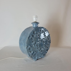 May include: A blue ceramic table lamp with a textured, spherical body. The lamp has a crackle glaze finish on one side and a raised, geometric pattern on the other. It features a white lamp holder and a white electrical cord.