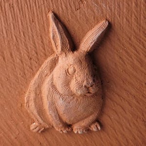 Puede incluir: Primer plano de una escultura en relieve de color terracota que representa un conejo. El conejo está representado en posición sentada con las orejas erguidas. La escultura tiene una superficie texturizada y los rasgos del conejo están claramente definidos.