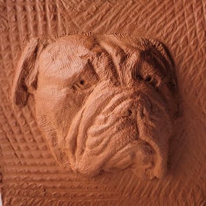 May include: A detailed, reddish-brown clay relief sculpture of a dog's head. The dog has a wrinkled face, droopy jowls, and a serious expression. The background has a textured pattern. The sculpture is likely handmade.