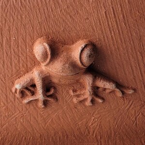 Peut inclure: Gros plan d'une sculpture de grenouille de couleur terre cuite. La grenouille est en relief, avec des détails en relief et une surface texturée. La grenouille a de grands yeux ronds et des membres tendus, ce qui lui donne une apparence réaliste.